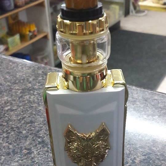 White and gold vape mod with wolf emblem. Glass tank on top. Sitting on countertop. Shelves visible in background.